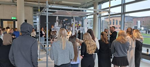 Eine große Gruppe von Studierenden stehen vor einer Installation aus Gerüststangen, die einen großen Tisch bilden, auf dem große  Architekturmodelle aus Pappe stehen. Von oben herab hängen eine Vielzahl an Bildern und handgeschriebenen Notizen.